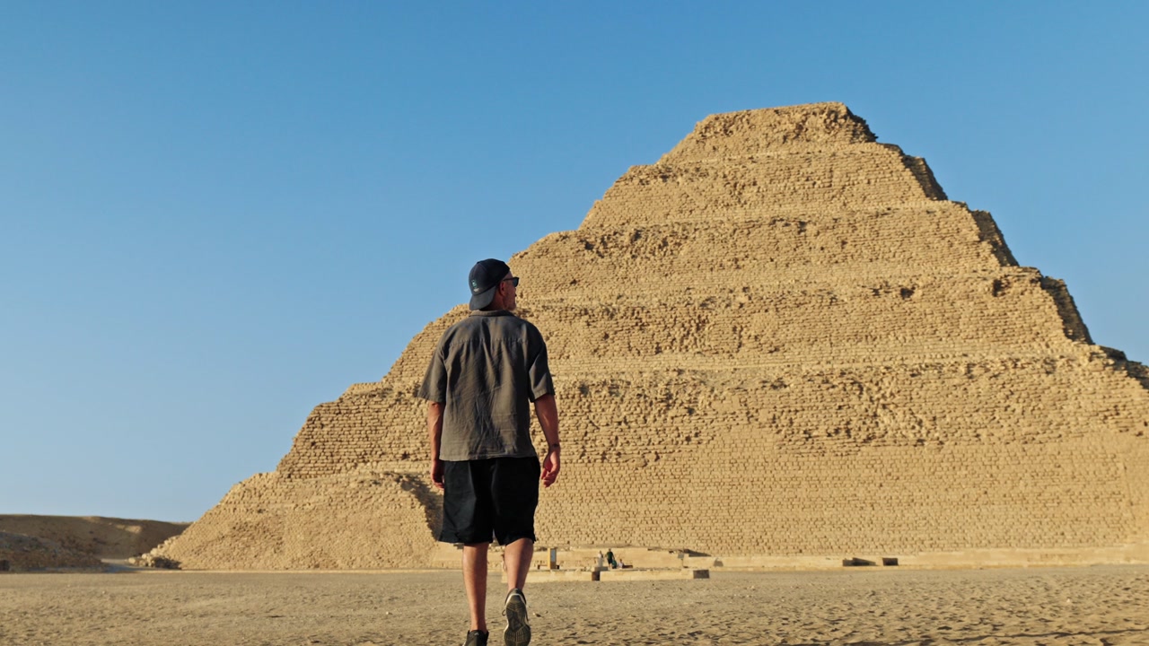 Man walking across the desert plateau of Saqqara as the sun sets, casting a warm, golden glow over the ancient landscape. In the background stands the iconic Step Pyramid of Djoser.