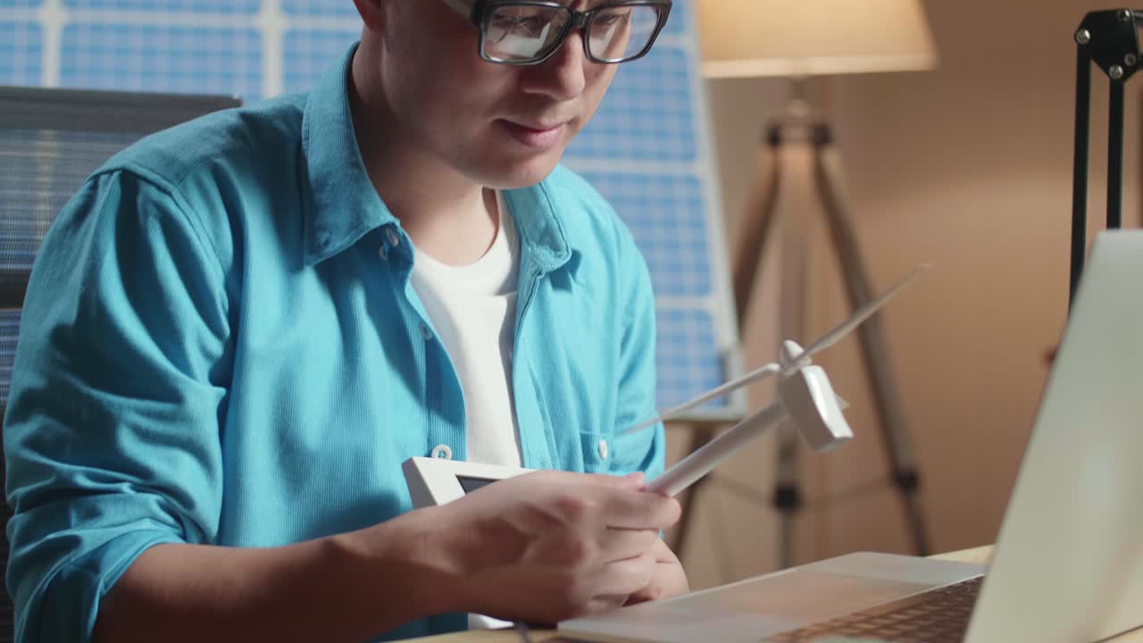 un hombre asiático sentado frente a una célula solar mira una turbina eólica mientras escribe en una computadora portátil en la oficina