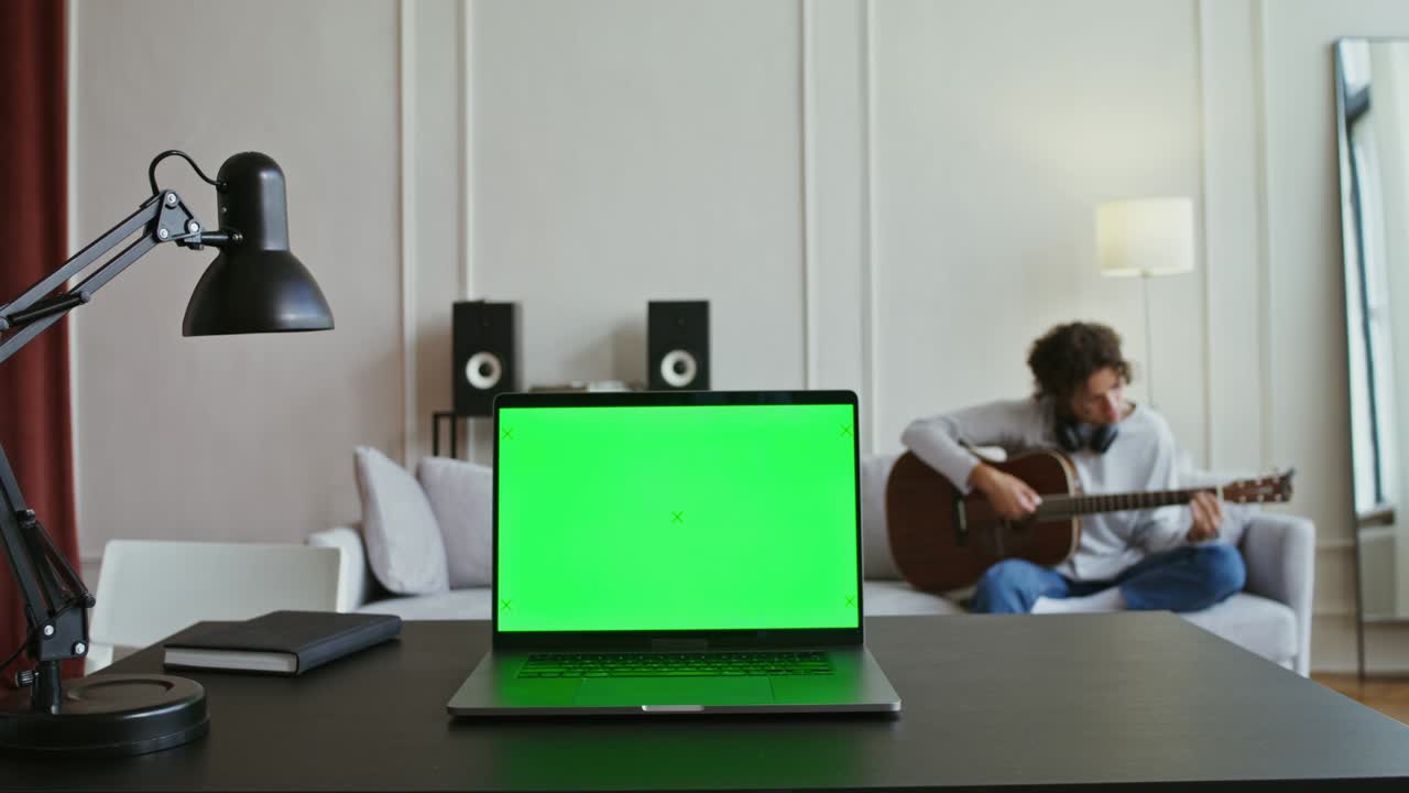A laptop with a green screen sits on table in front of a man playing guitar