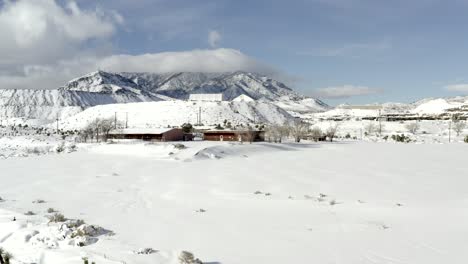 Aerial-view-rising-above-winter-chalet-cabin,-snow-covered-landscape-mountain-peaks-in-far-distant-background