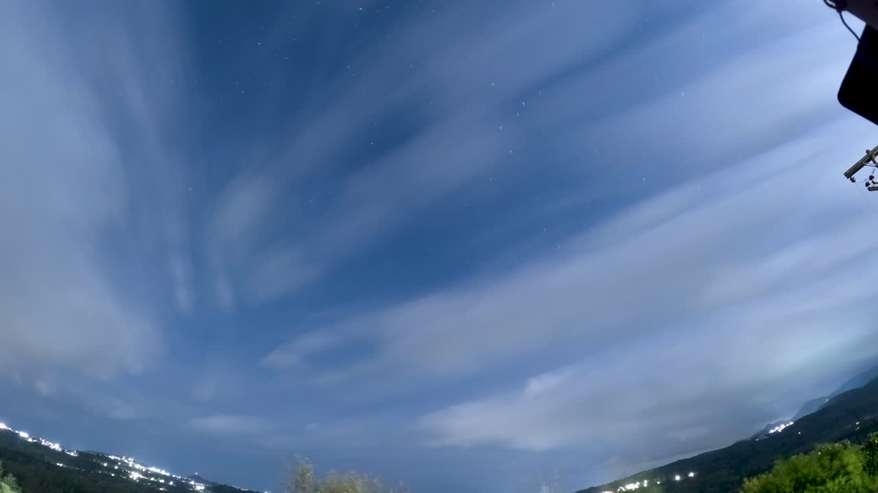Time lapse grandangolare di un cielo notturno blu con nuvole filiformi in movimento e stelle visibili sopra un paesaggio rurale con luci della città in lontananza a Corfù, in Grecia