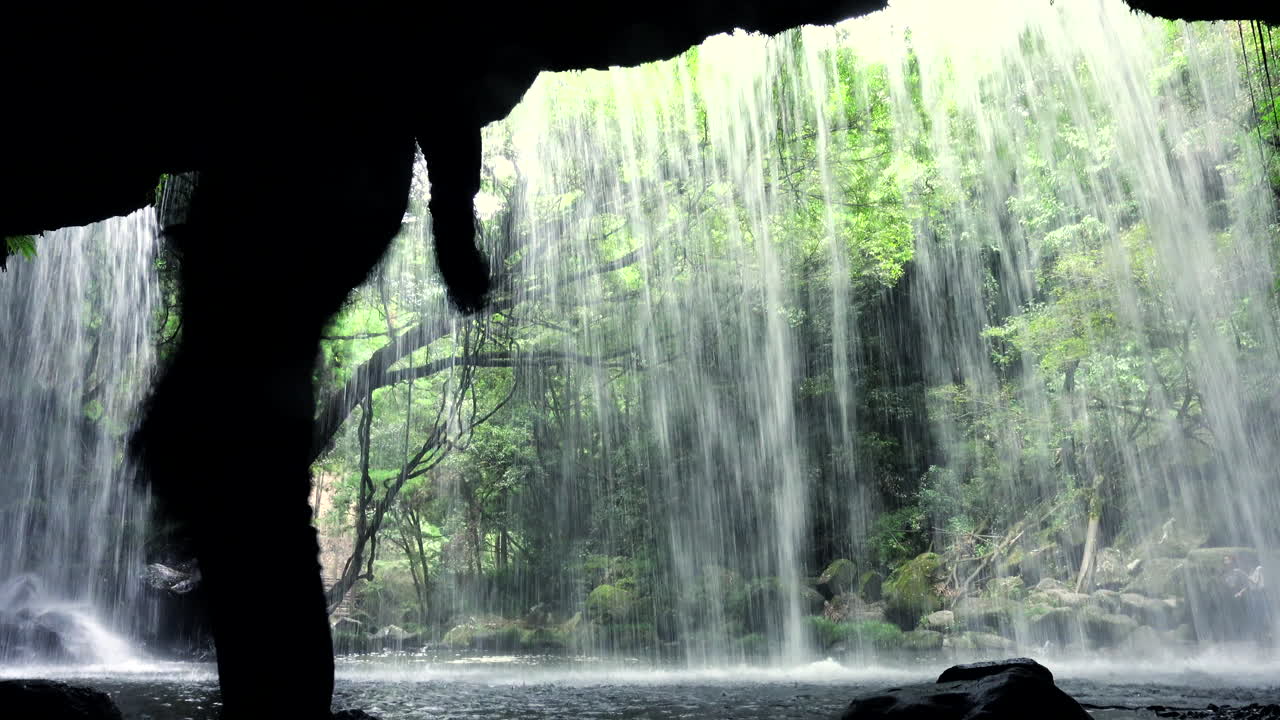 Silhouettes of people crossing in front of the camera behind an impressive waterfall in the tourist Nabegataki Falls Kumamoto Japan green nature. landscape back view shot