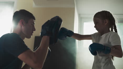 master and kid sparring at friendlies in gym. professional trainer uses hand pads to exercises little girl in sports club slow motion. combat lesson