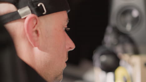 close-up of a man s face wearing a black cap, with a focused expression. in the blurred background, a speaker is visible, indicating a studio or audio production setting