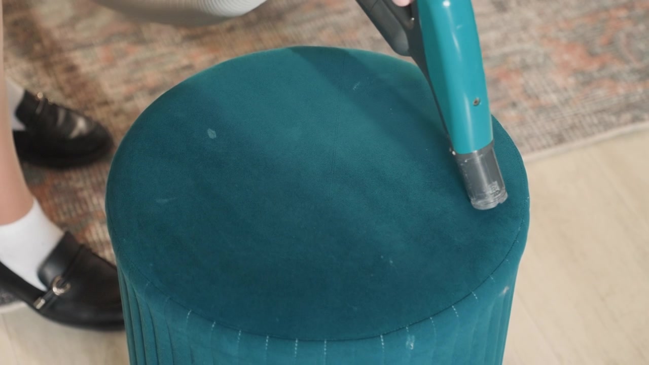 Pouf color verde acqua aspirato in soggiorno accanto a scarpe visibili, l'ugello portatile si muove sulla pila di tessuto rimuovendo polvere e detriti, piedi e calzini indicano il proprietario di casa che si occupa delle faccende domestiche, rapida routine di pulizia