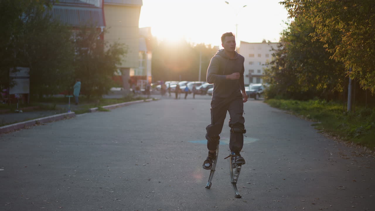 회색 후디와 어두운 회색 바지를 입은 짧은 갈색 머리카락을 가진 사람이 주거 지역의 포장 된 도로에서 스프링 스트를 사용합니다. 먼 곳에서 잡혔고, 배경에는 나무와 건물이 있습니다.