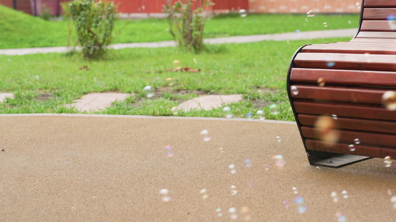 Close up of colorful soap bubbles drifting over park path with wooden bench on right, soft focus greenery and lawn behind, gentle bokeh highlights, calm outdoor vibe