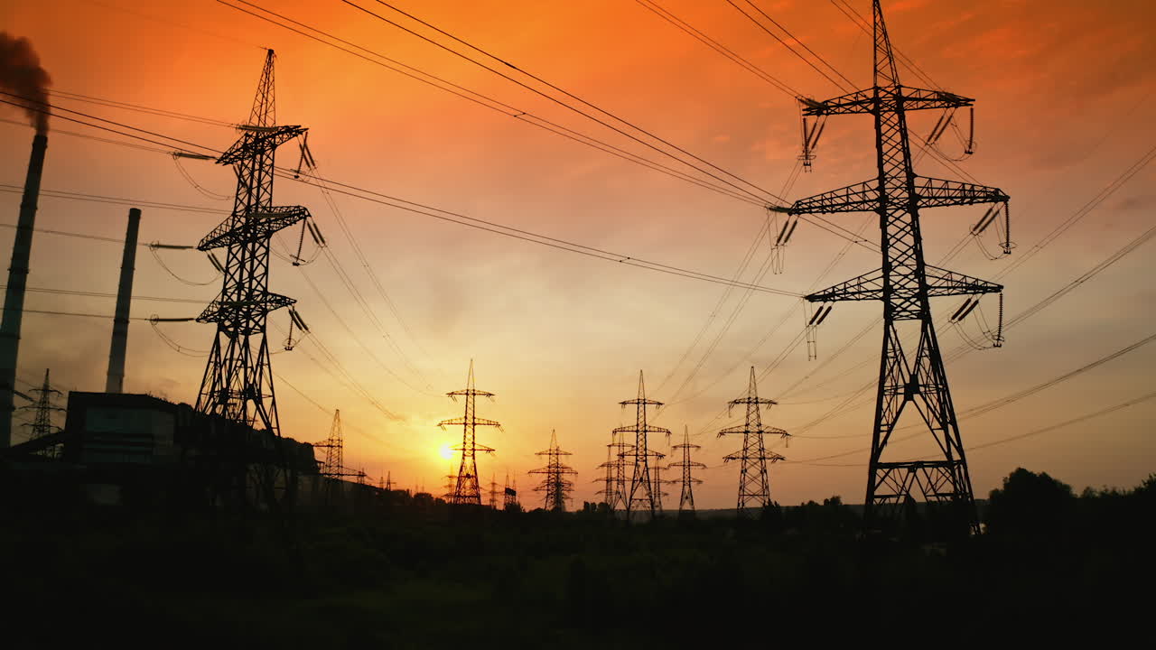 Electric towers against orange sky at sunset. High-voltage electricity pylons in the evening. Panoramic view of transmission lines. Camera rising.
