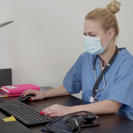 Confident-experienced-young-doctor-woman-with-face-mask-sitting-in-front-of-computer-in-office