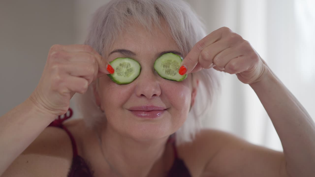 Cheerful woman having fun taking care of facial skin at home indoors. Portrait of positive Caucasian lady with cucumber circles closing opening eyes looking at camera. Femininity and domestic routine.