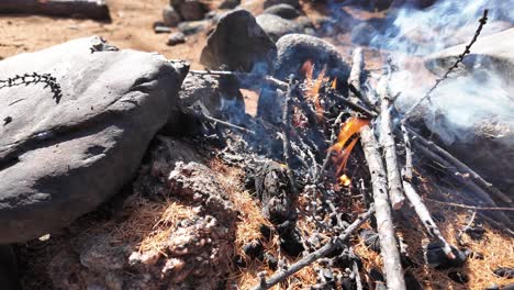 A-small-campfire-crackling-between-large-rocks-in-a-forest-area-with-autumn-leaves-scattered-around,-emitting-smoke-and-small-flames