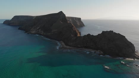 Impresionante-Vuelo-Panorámico-De-Verano-Sobre-Las-Olas-Del-Mar-Del-Océano-Turquesa-Rodando-En-La-Playa-De-Arena-De-Calheta-Hacia-El-Pintoresco-Islote-De-Cal-Espectacular-Isla-Marrón-Y-Pico-De-La-Montaña-Al-Atardecer,-Enfoque-Aéreo