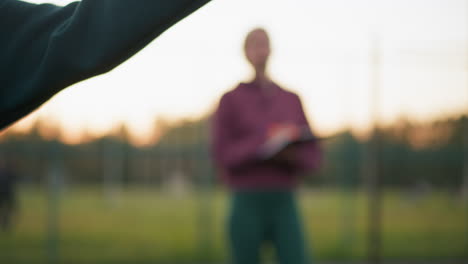 close-up of volleyball held by athlete in green sweater, preparing to serve, with blurred background featuring coach observing and people walking across field in outdoor training session