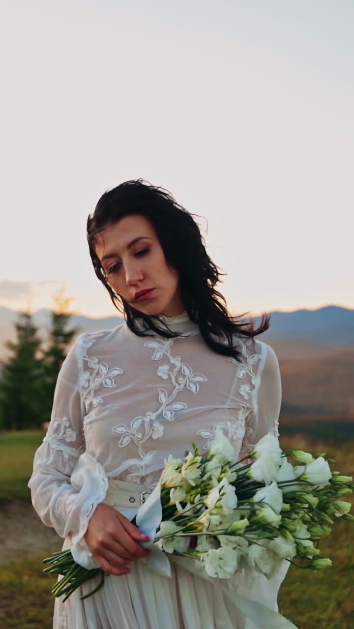 Lonely woman in white dress walks closer to the camera holding bouquet in her hands. Wind fluttering lady's hair and dress. Amazing mountain landscape at setting sun at backdrop. Vertical video