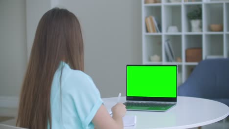 niña tomando notas en el bloc de notas de la computadora portátil pantalla de maqueta verde. niña aprendiendo en línea en la computadora