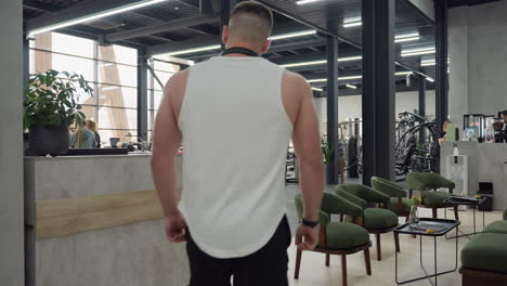 well bodied man walks into modern gym through reception hall as people exercise on equipment, weights, cardio machines and free training areas under bright natural light from large glass windows