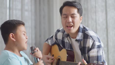 close up of asian father and son with expressive face enjoys singing and playing the guitar together on sofa at home