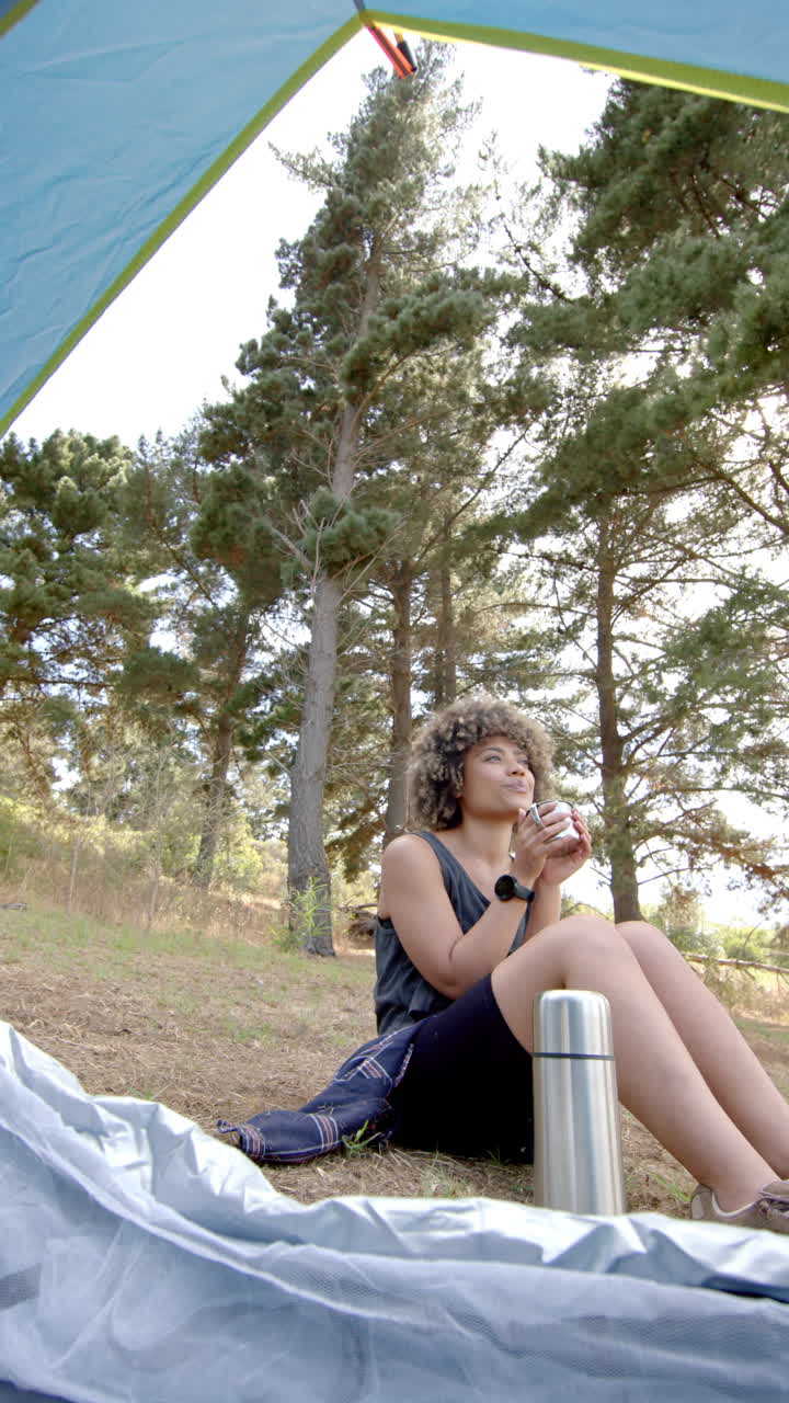 Vertical video: Camping in forest, woman enjoying hot drink outside tent with thermos nearby