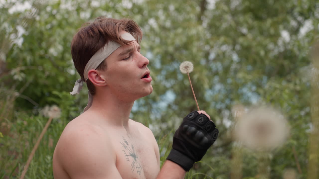 Adventurer wearing gloves and headband holds delicate dandelion stem, blowing seeds into breeze while standing shirtless in lush forest, expression focused, surrounded by green foliage and summer calm