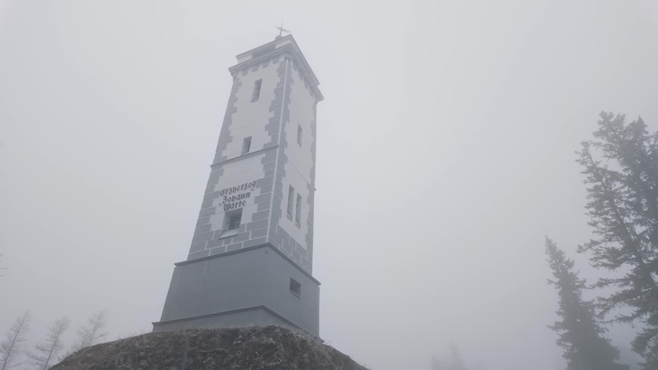 The Erzherzog Johann tower in Mariazell is shrouded in fog, standing tall amidst the cold, wintery landscape. This iconic landmark is surrounded by towering trees and a frosty atmosphere.
