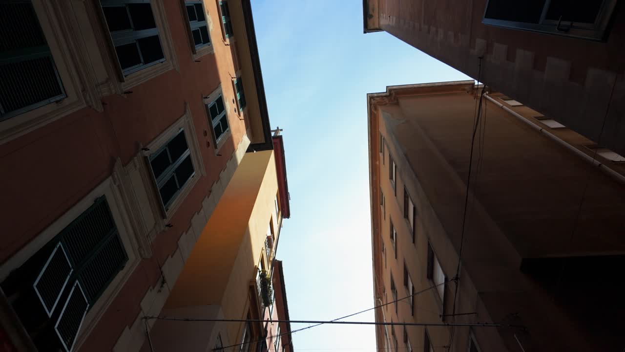 Drammatica inquadratura grandangolare che guarda in alto tra alti palazzi color ocra con finestre aperte, che proiettano ombre, sotto un cielo azzurro brillante. Geometria urbana, luce naturale, nessuna persona