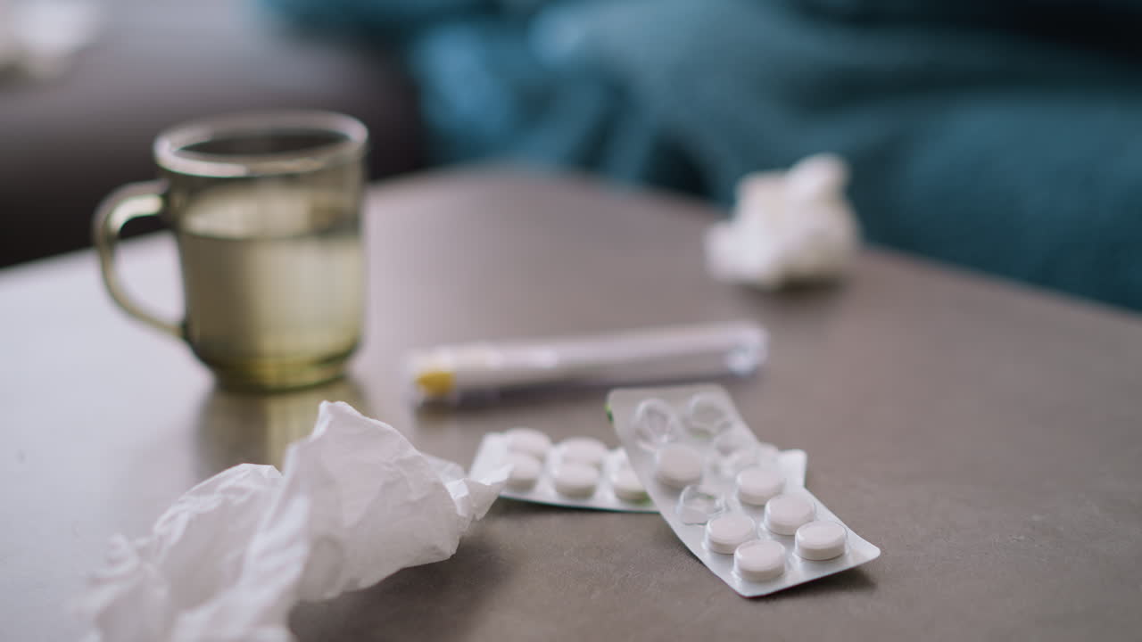 Close-up of drugs, used tissue, glass cup, and thermometer on table with light reflections, indicating illness recovery, showing signs of flu or cold, with a focus on medication and symptoms
