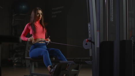 the girl performs an exercise on the muscles of the back. she sits behind a special training machine pulls the handle to the lower abdomen and returns to its original position.