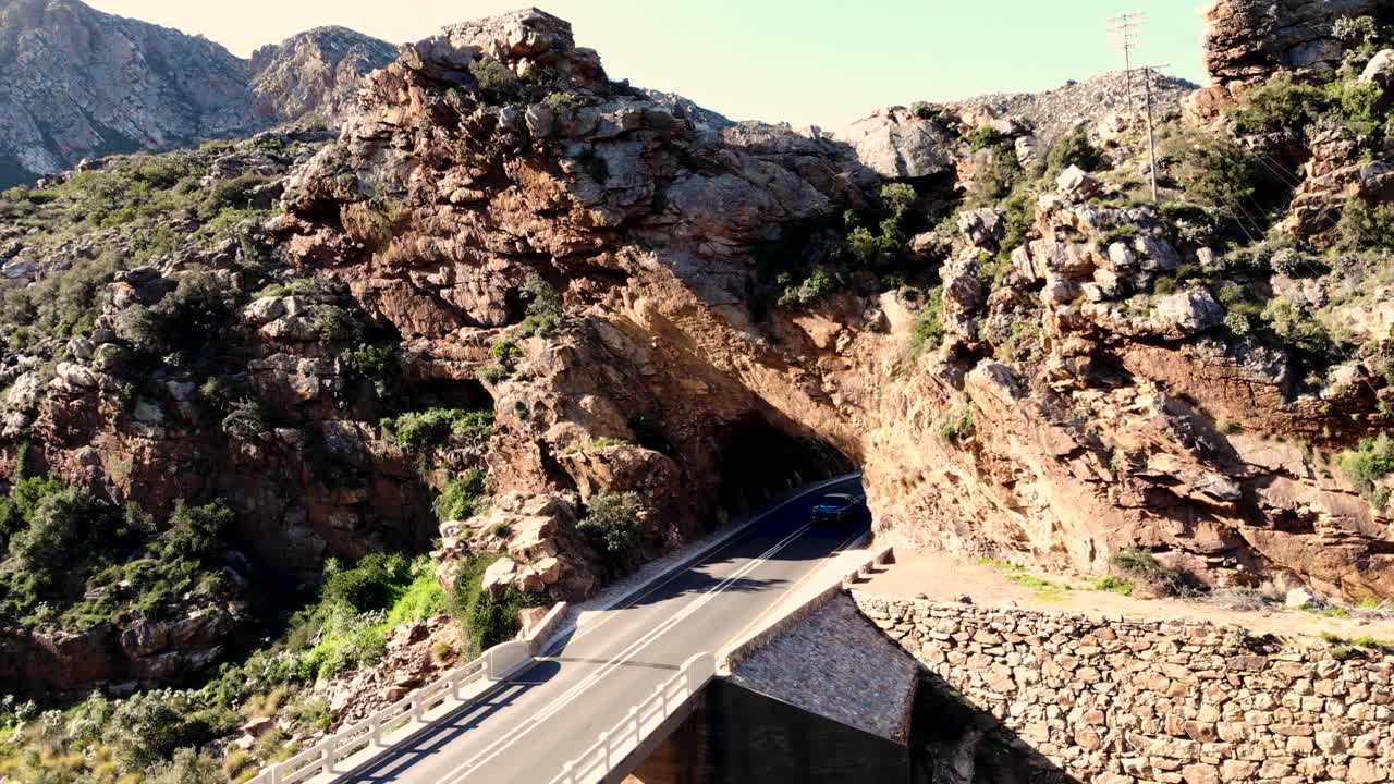 A captivating drone's-eye view captures a blue car journeying through a mountain tunnel, emerging into the radiant light beyond, symbolizing adventure and exploration