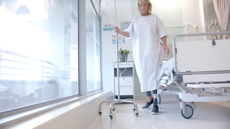 kaukasische ältere patientin mit prosthetischem bein geht im krankenhaus und hält einen iv-stand