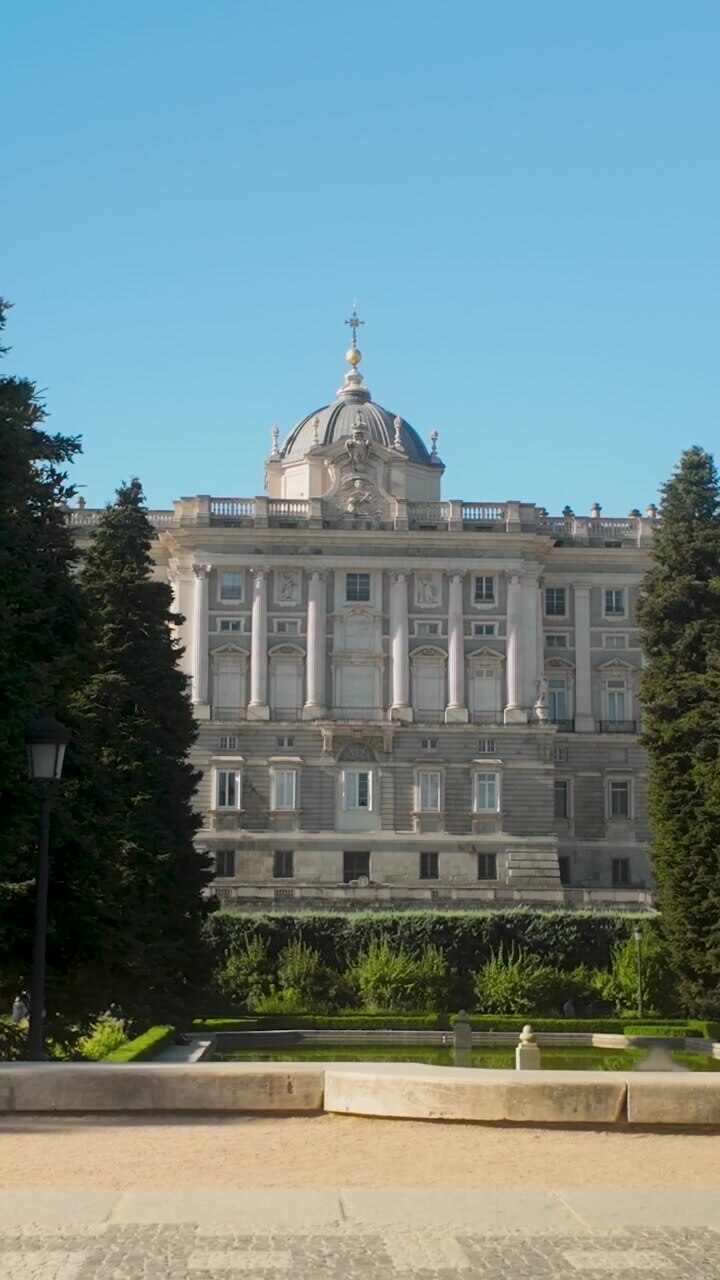Una prospettiva verticale dell'iconico Palazzo Reale di Madrid, incorniciato da alberi verdi lussureggianti nei Giardini di Sabatini. Adatto per documentari di viaggio e contenuti sul turismo spagnolo