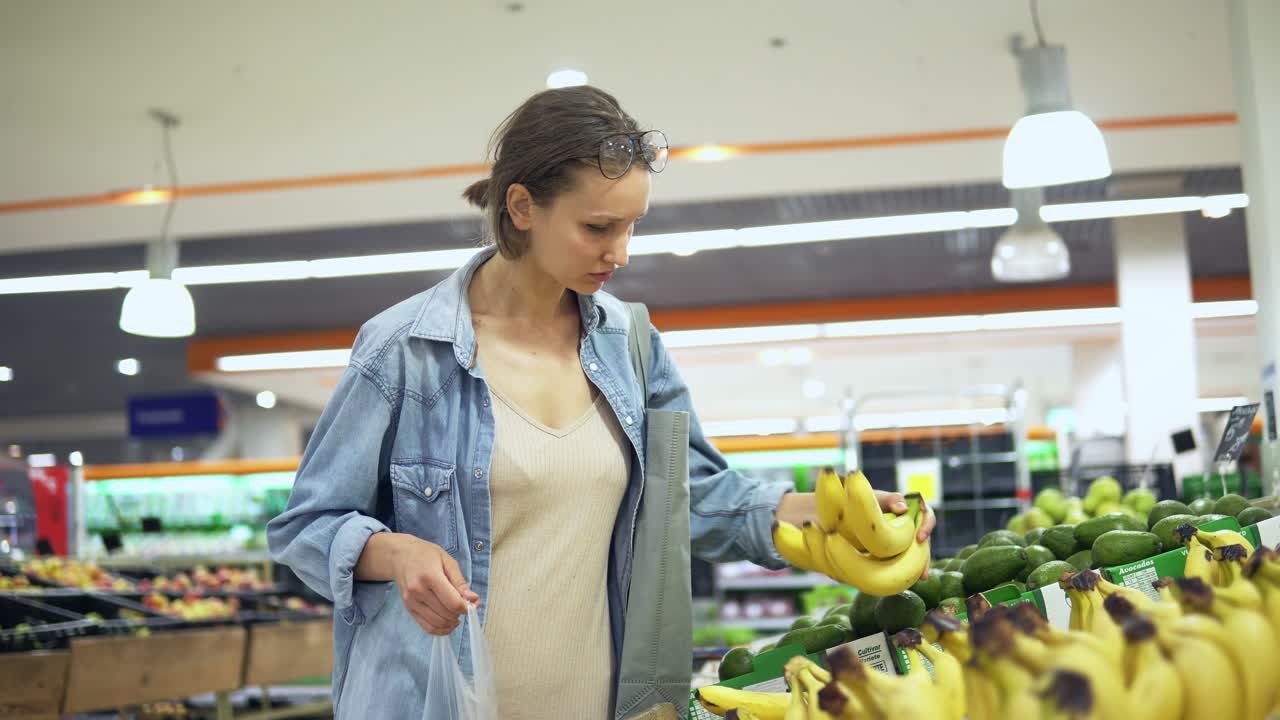 imágenes de mano de una chica joven y bonita en camisa seleccionando frutas en el supermercado. pone algunos plátanos en una bolsa de celofán. concentrado. estilo casual