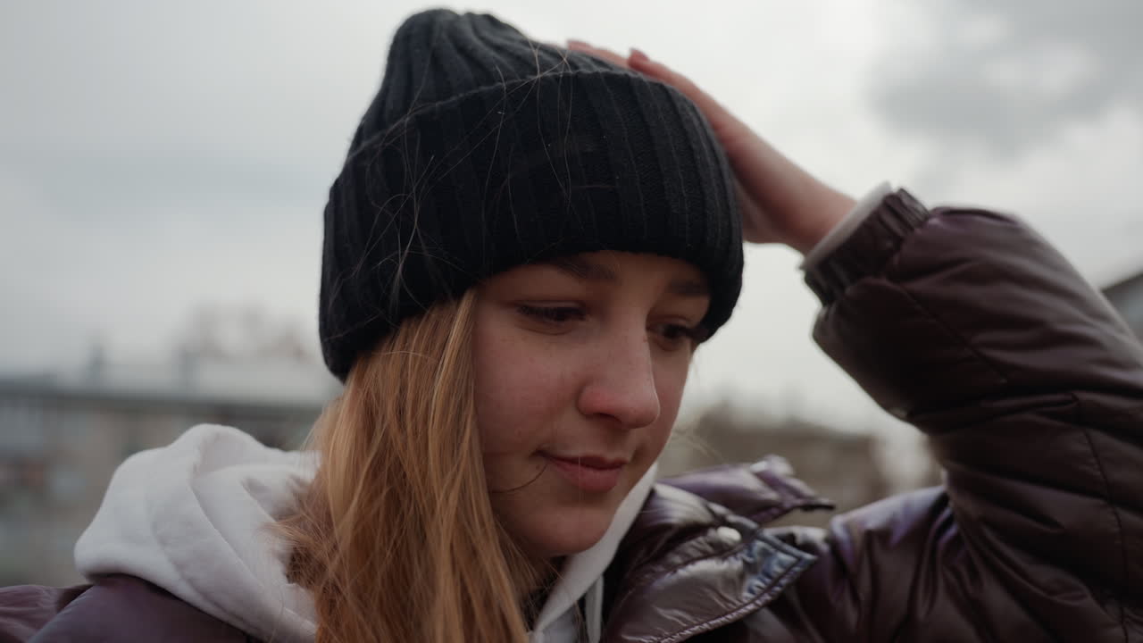 비니, 후드티, 패딩 코트를 입고 야외에 있는 젊은 백인 여성, 흐린 하늘과 아파트 배경으로 클로즈업하여 자연스럽게 촬영, 아래를 향한 시선, 부드러운 자연광, 조용한 겨울 분위기, 통근하는 학생