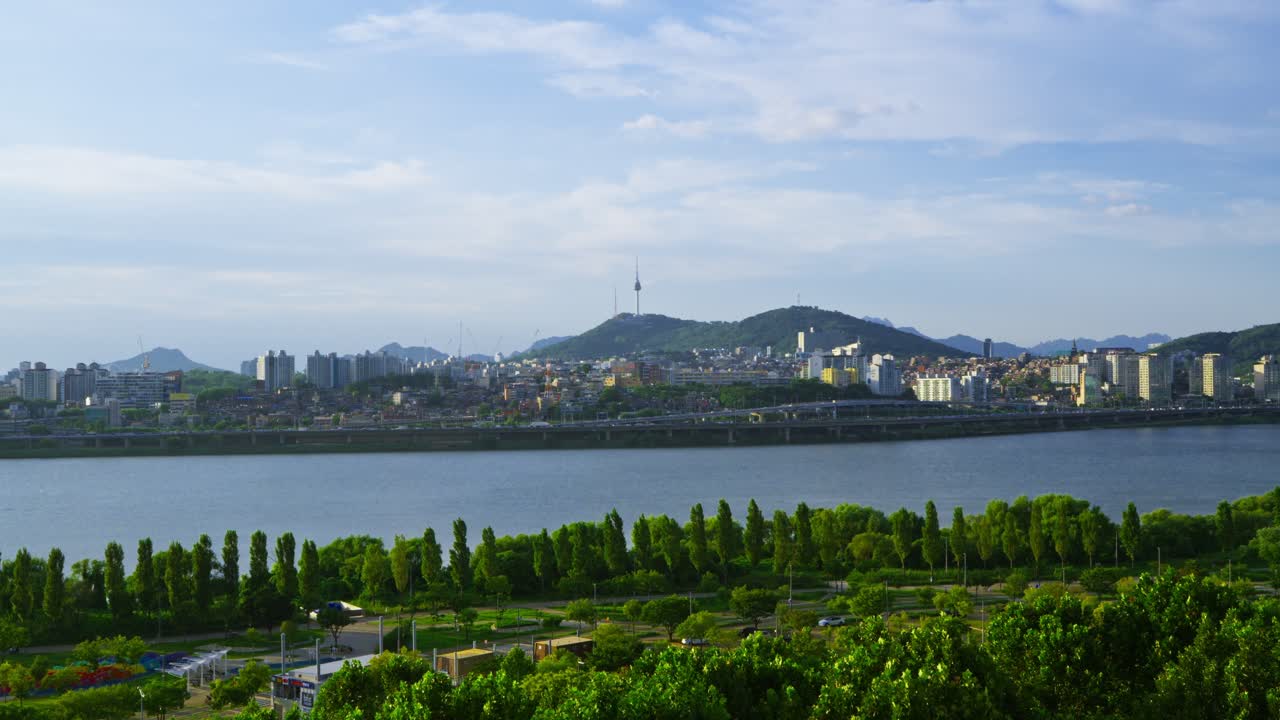 Spacious aerial view of Seoul showing green Han River park in the foreground with Yongsan district skyline and N Seoul Tower on a cloudy summer day