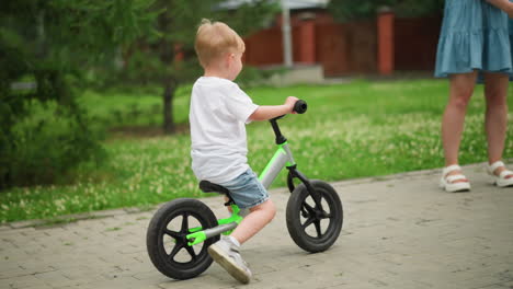 a joyful little boy rides his bicycle on a paved walkway, heading towards a woman standing by the side