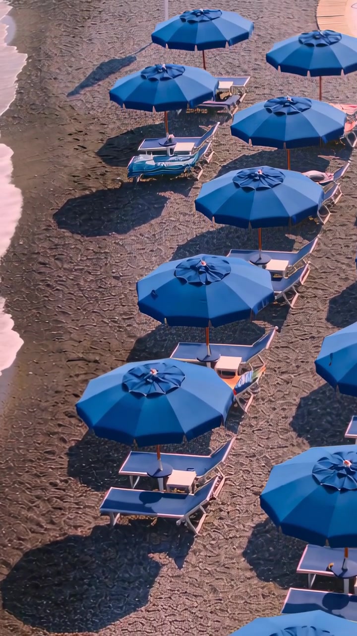 Video verticale: Telecamera panoramica che rivela una fila di ombrelloni blu e lettini vuoti sulla spiaggia, con un'onda.