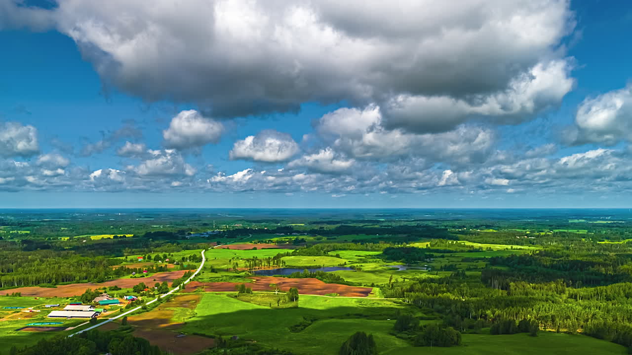A stunning forward-moving aerial hyperlapse glides over a vast and picturesque Latvian landscape of agricultural fields and dense forests, with scattered white clouds moving rapidly across the sky