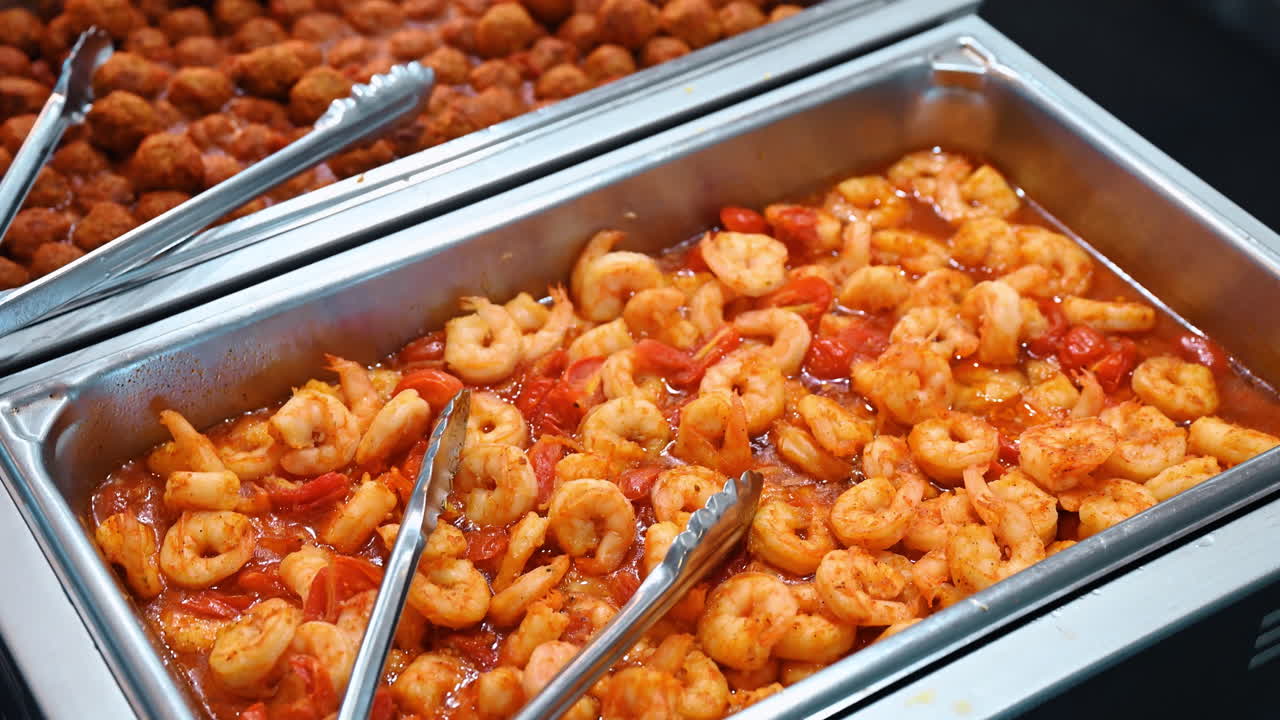 Gamberi cotti in salsa di pomodoro in vassoio metallico da buffet. Chafing dish in acciaio inossidabile riempito con gamberi sgusciati in salsa rossa di pomodoro e aglio con pinze da portata in metallo