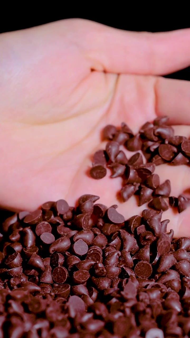 Una mano si immerge in un mucchio di gocce di cioccolato fondente, raccogliendole e setacciandole.