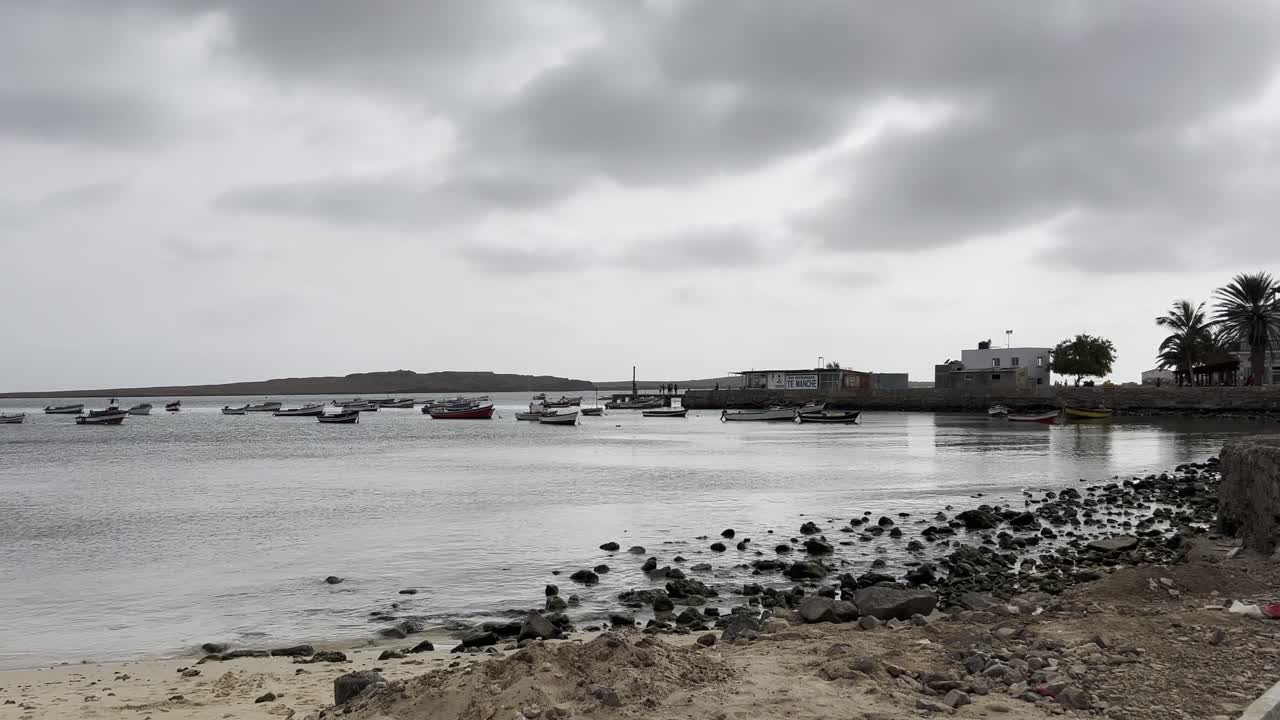 Serene view of Sal Rei harbor in Cape Verde featuring moored fishing boats, a rocky shoreline, and overcast skies creating a tranquil atmosphere