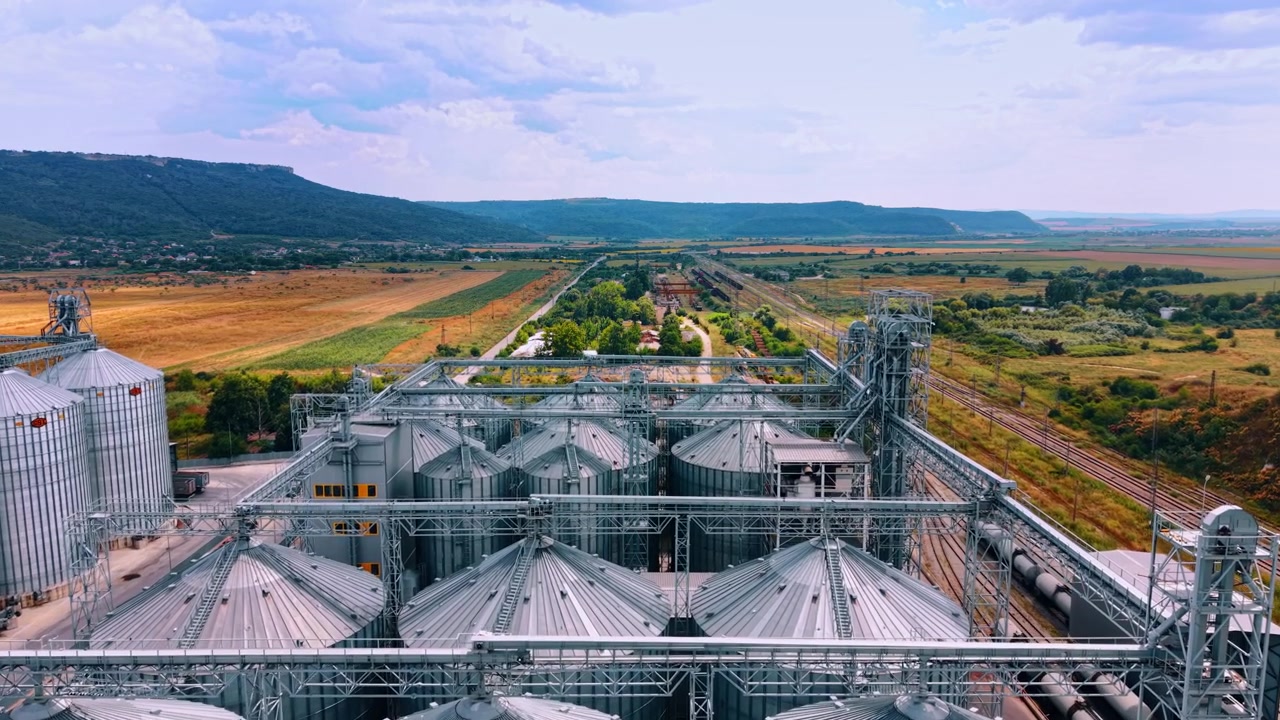 Varna, Bulgaria, 3 Jule 2025 : Aerial panorama of grain silo facility near countryside. Metal storage towers overlook distant green hills and agricultural fields under a cloudy sky.