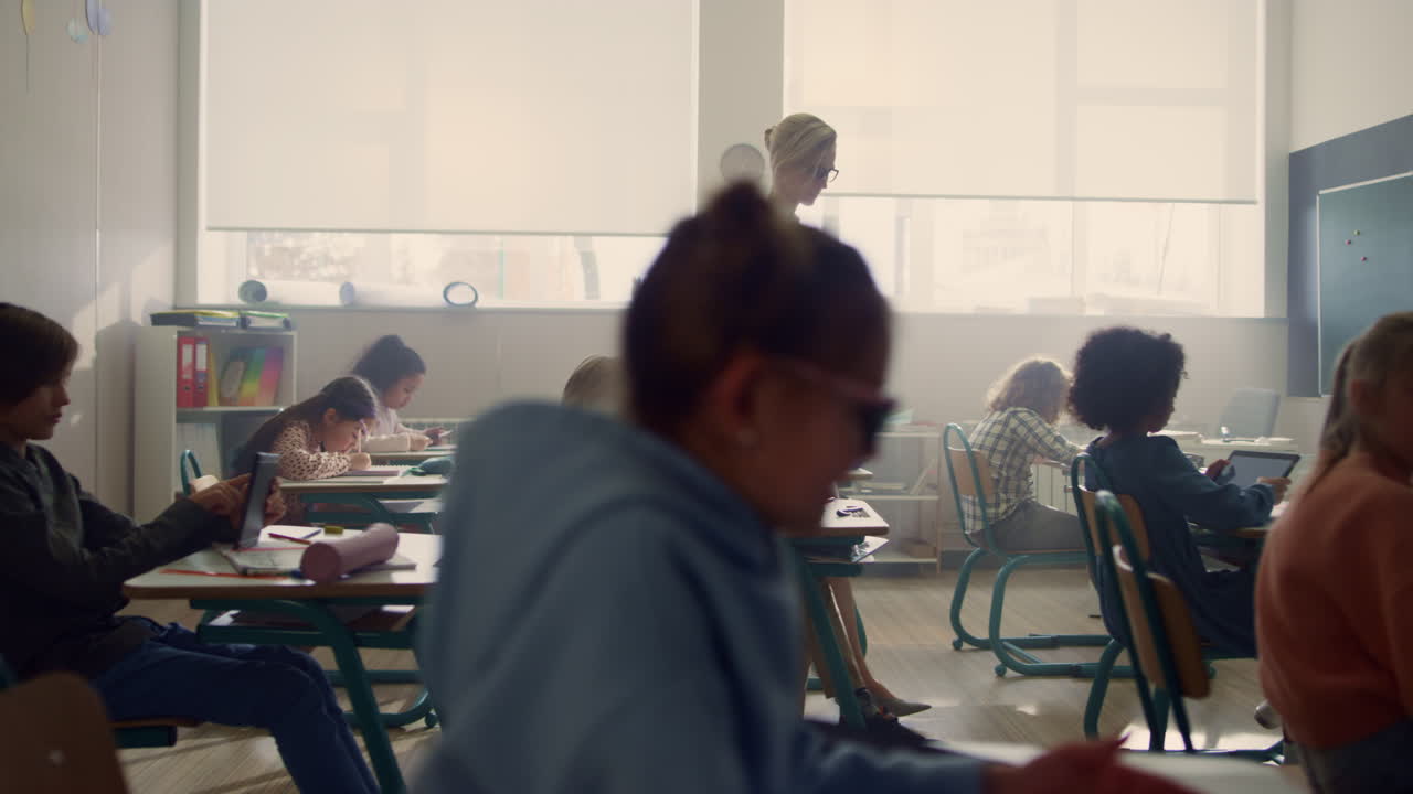 Schoolteacher walking in classroom with students. Pupils doing test on tablets