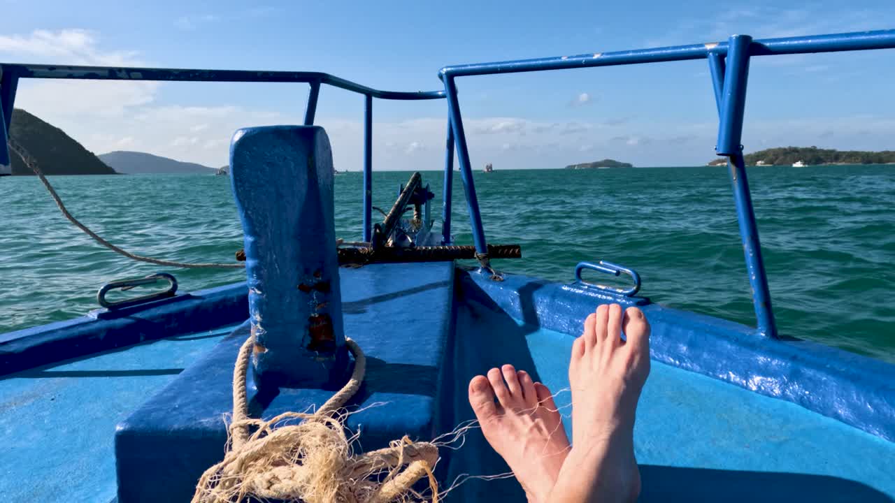 Feet rest on a blue boat's deck, cruising through Phuket's vibrant sea under clear skies, capturing a serene, leisurely moment