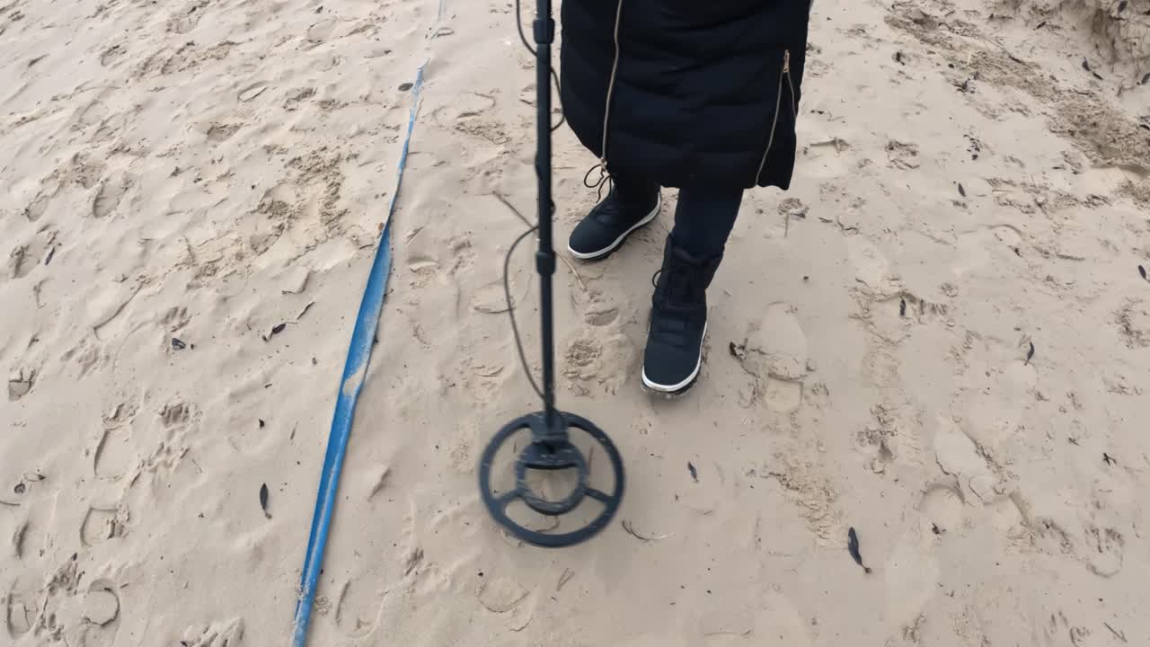 Persona che esamina un terreno sabbioso con un metal detector accanto a una lunga striscia blu distesa sulla spiaggia, muovendosi lentamente su impronte e detriti sparsi