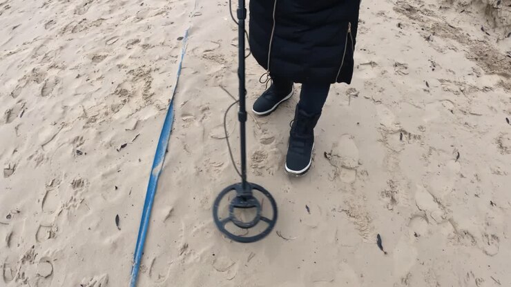 Persona che esamina un terreno sabbioso con un metal detector accanto a una lunga striscia blu distesa sulla spiaggia, muovendosi lentamente su impronte e detriti sparsi