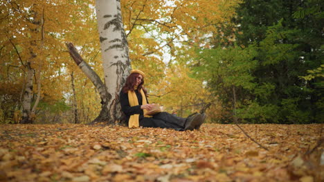 stylish lady in yellow beret relaxes outdoors, leaning against tree while flipping through book, surrounded by golden autumn leaves scattered on ground, enjoying peaceful seasonal atmosphere