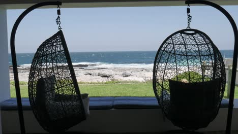 Sillas-Colgantes-En-La-Veranda-Que-Se-Balancean-Suavemente-En-La-Brisa-Del-Océano-Con-Hermosas-Vistas-Al-Océano-En-El-Fondo-En-Hermanus,-Sudáfrica