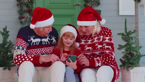 Abuelos-De-La-Familia-Mayor,-Nieta-Compran-Regalos-De-Navidad-En-Línea-En-Teléfonos-Móviles,-Compras