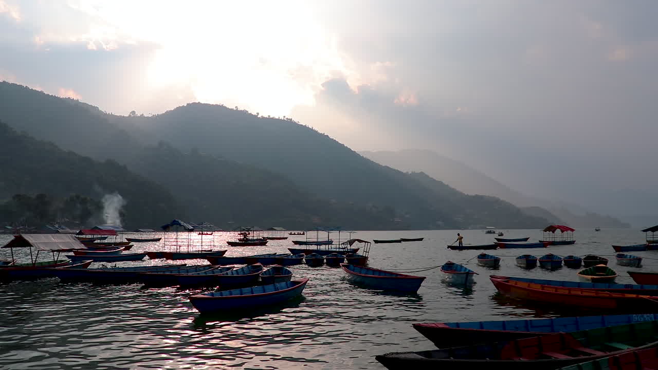 Traditional Wooden Boats at Phewa Lake in Pokhara, Nepal on a fine cloudy evening. SIlhouetted person rows the boat in background.
