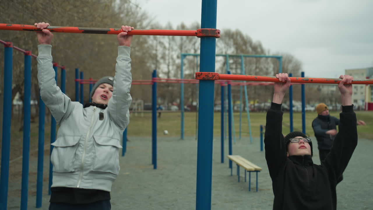 Two boys are working out on pull-up bars in an outdoor gym area, one boy is actively doing a pull-up, looking tired, while the other boy joined him on the adjacent bar