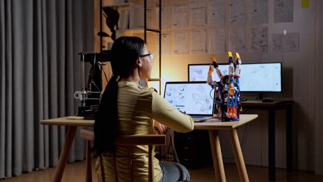 vista trasera de una adolescente asiática con impresión 3d comprobando una mano de cyborg mientras la diseña en una computadora portátil en casa